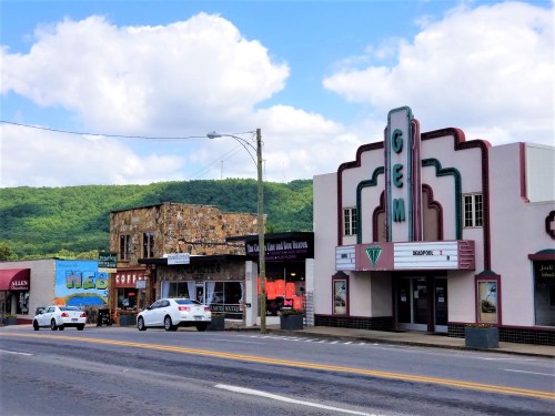 A street view featuring a vintage theater, shops, and green hills under a partly cloudy sky.