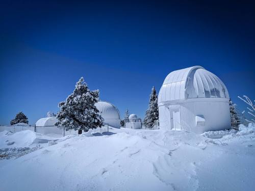 Snow-covered observatories surrounded by trees under a clear blue sky.
