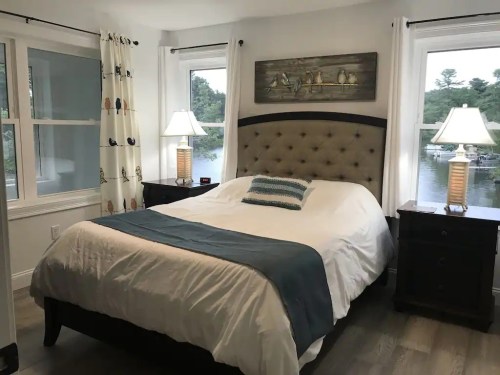 Cozy bedroom with a tufted headboard, two lamps, and windows overlooking a serene lake.