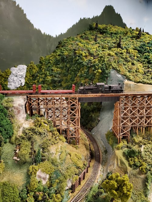 A model train crosses a wooden trestle bridge over a scenic landscape of lush greenery and hills.