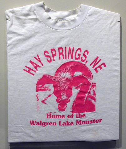 White t-shirt featuring a pink graphic of a monster and text: "Hay Springs, NE Home of the Walgren Lake Monster."