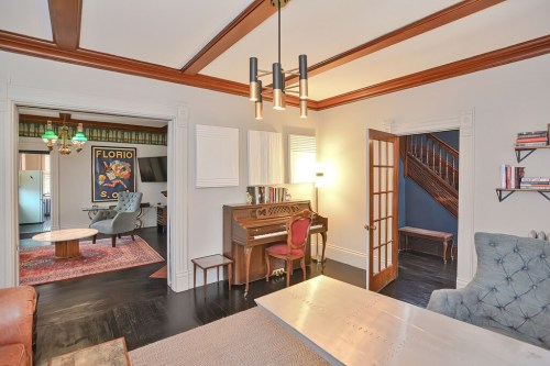 Cozy living room with a piano, vintage decor, and a view of a staircase and adjoining room. Warm, inviting atmosphere.