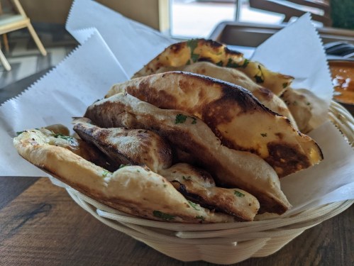 A basket of freshly baked naan bread, lightly charred and garnished with herbs, resting on parchment paper.