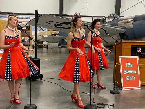 Three women in red dresses with star patterns perform on stage, singing into microphones in an aircraft hangar.