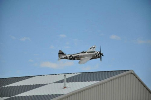 A vintage silver airplane with a checkered tail flying above a building under a blue sky.