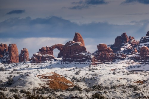 Snow-covered red rock formations under a cloudy sky, featuring a prominent arch in the landscape.