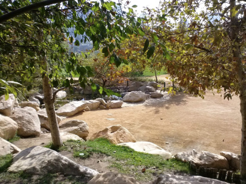 A serene outdoor scene with large rocks, green grass, and trees, overlooking a calm area with a small pond.