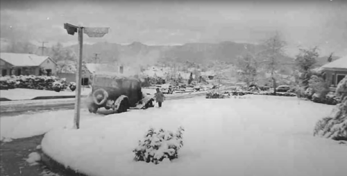 A snowy street scene with a vintage car, houses, and mountains in the background, creating a serene winter landscape.