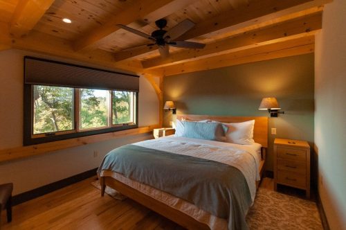 Cozy bedroom with a wooden ceiling, a queen bed, bedside lamps, and a large window overlooking greenery.