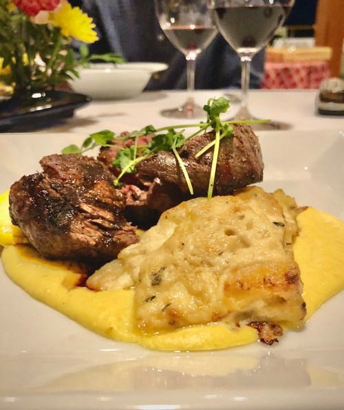 A plate of tender beef with herbs, served on creamy polenta, accompanied by a side of rich, cheesy dish. Wine glasses in background.