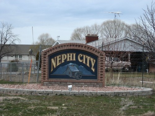 Sign for Nephi City featuring a wagon and mountains, set in a grassy area with a cloudy sky in the background.