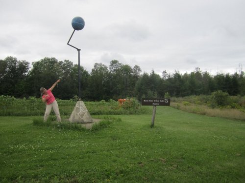 A person in a pink shirt leans against a tall sculpture of a globe on a pole in a grassy area with trees in the background.