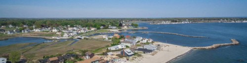 Aerial view of a coastal town with houses, a sandy beach, and calm blue waters under a clear sky.