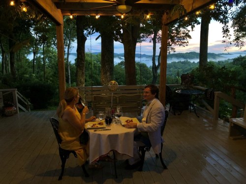 A couple dining at a table on a porch, surrounded by trees, with a scenic sunset view in the background.