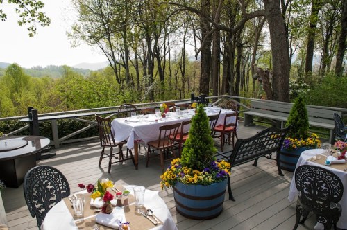 A scenic outdoor dining area with tables set among trees and colorful flowers, overlooking a lush green landscape.