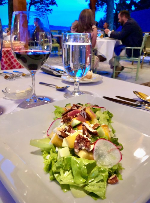 A fresh salad with greens, fruits, nuts, and radishes, set on a dining table with wine and water glasses in the background.