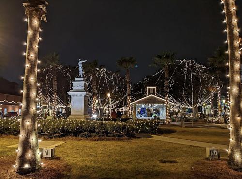 A festive park at night, adorned with lights, featuring a statue and a small decorated building among palm trees.