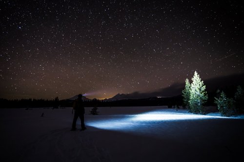 A person stands in a snowy landscape at night, illuminated by a flashlight, with a starry sky and mountains in the background.