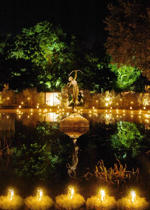 A serene night scene featuring a statue by a pond, illuminated by soft lights and surrounded by lush greenery.