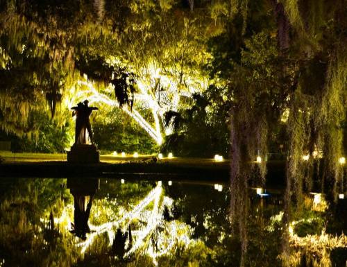 A serene night scene featuring a statue surrounded by illuminated trees, reflecting in a calm pond.