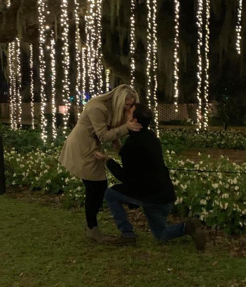 A couple shares a romantic kiss in a garden adorned with twinkling lights and lush greenery.