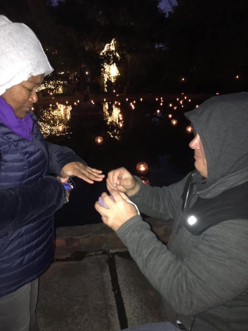 A person kneels by a pond, proposing to someone wearing a jacket and a hood, surrounded by glowing lights.