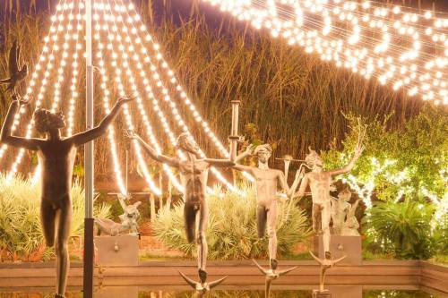 A garden scene featuring illuminated sculptures of figures holding hands, surrounded by lush greenery and string lights.