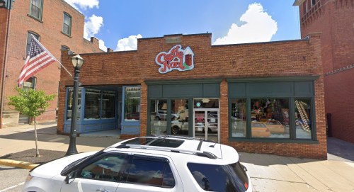 A brick storefront with a colorful sign, featuring large windows and an American flag nearby. A white SUV is parked in front.