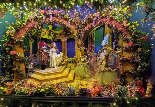 A whimsical display featuring figures among vibrant flowers and greenery, set in an ornate, fairy-tale-like scene.