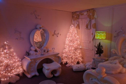 A whimsical room with pink walls, two lit Christmas trees, a stylish vanity, and a retro lamp, creating a cozy atmosphere.