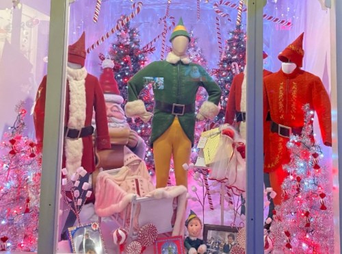 A festive display featuring elf costumes, Santa outfits, and decorated Christmas trees with colorful lights.