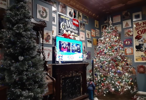 Cozy room decorated for Christmas, featuring two trees, a TV displaying holiday content, and festive wall art.