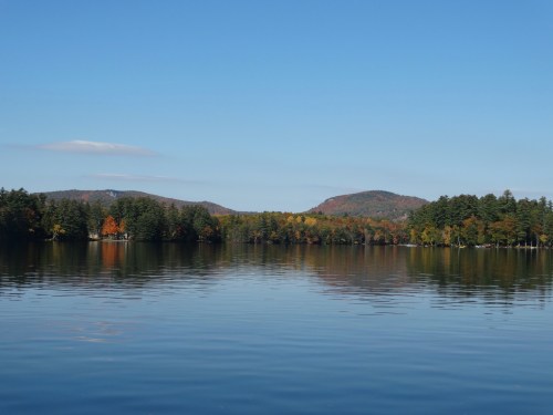 A serene lake surrounded by colorful autumn trees and distant hills under a clear blue sky.