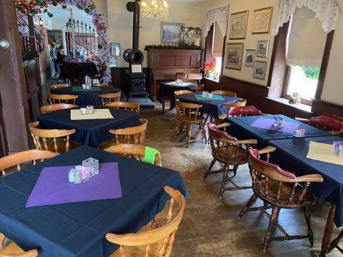 Cozy dining area with wooden tables, purple and beige tablecloths, and a vintage piano in a warmly lit room.