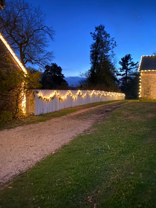 A gravel path lined with festive lights along a white fence, surrounded by trees at dusk.