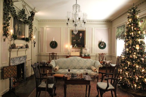 A cozy, elegantly decorated room with a Christmas tree, fireplace, and vintage furniture, adorned with festive wreaths.