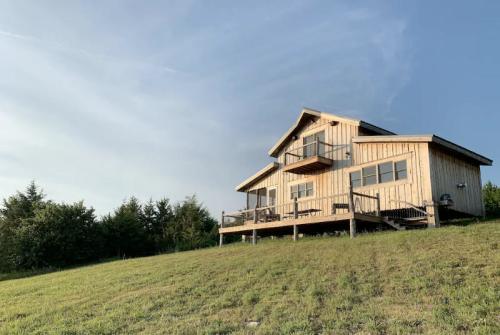 A wooden house on a grassy hill, featuring a deck and large windows, under a clear blue sky.