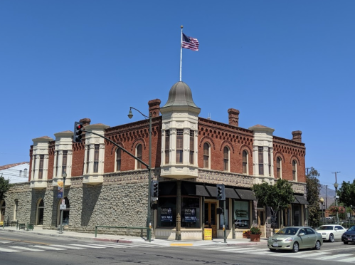 Historic brick building with a dome and flag, featuring large windows and a corner storefront, against a clear blue sky.