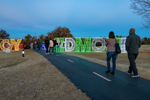 People walking along a path past large, illuminated letters spelling "JOY" and "EDMOND" against a twilight sky.