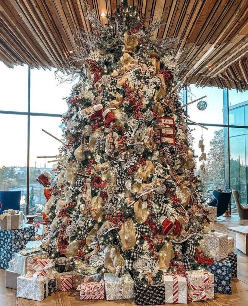 A beautifully decorated Christmas tree with gifts underneath, featuring red, gold, and black patterns, in a bright room.