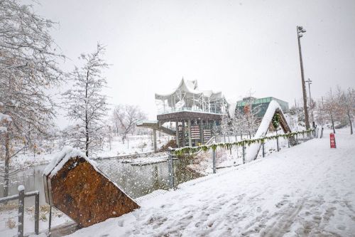 A snowy landscape featuring a modern building, trees, and a pathway along a frozen body of water.