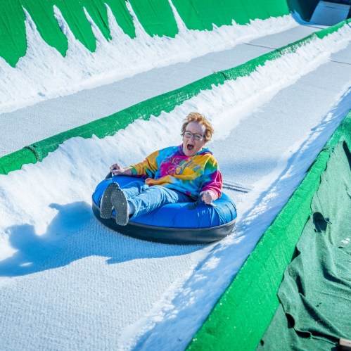A child in a colorful tie-dye shirt joyfully sleds down a snowy slope on a blue inner tube.