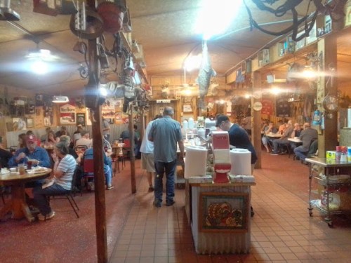 A cozy restaurant interior with diners at tables, a counter with food, and rustic decor hanging from the ceiling.