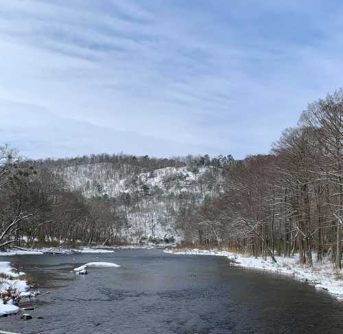 A serene winter landscape featuring a river surrounded by snow-covered trees and distant hills under a cloudy sky.