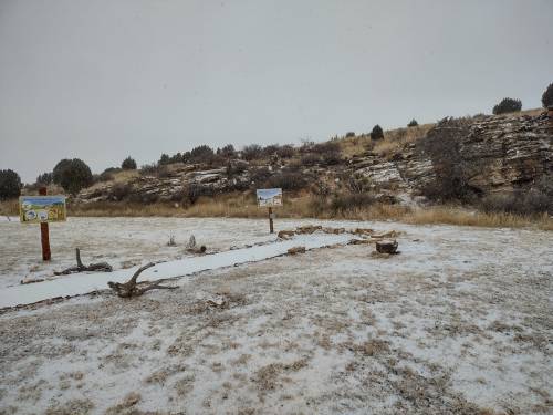 A snowy landscape with signs and rocky hills in the background, surrounded by sparse vegetation.