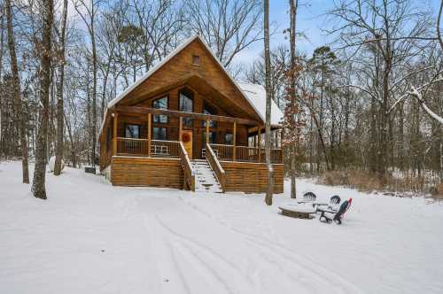 A cozy wooden cabin surrounded by snow-covered trees, featuring a front porch and a fire pit area.