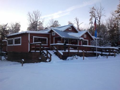 A red house with a porch, surrounded by snow and trees, under a clear sky. An American flag is visible.