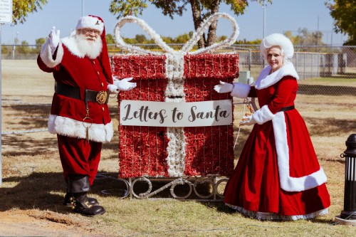 Santa and Mrs. Claus stand beside a large, festive gift box labeled "Letters to Santa" in a sunny outdoor setting.