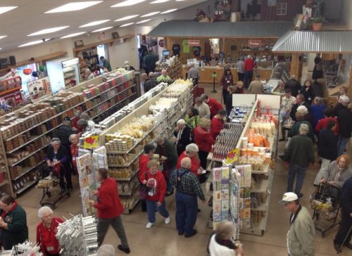 A busy store interior filled with shoppers browsing aisles of products, with many people in red jackets.