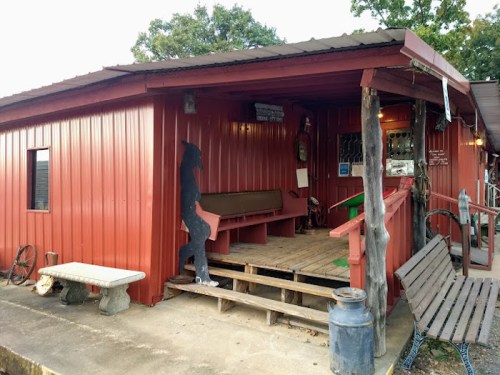 A red wooden building with a porch, featuring a bench and a silhouette figure standing by the entrance.